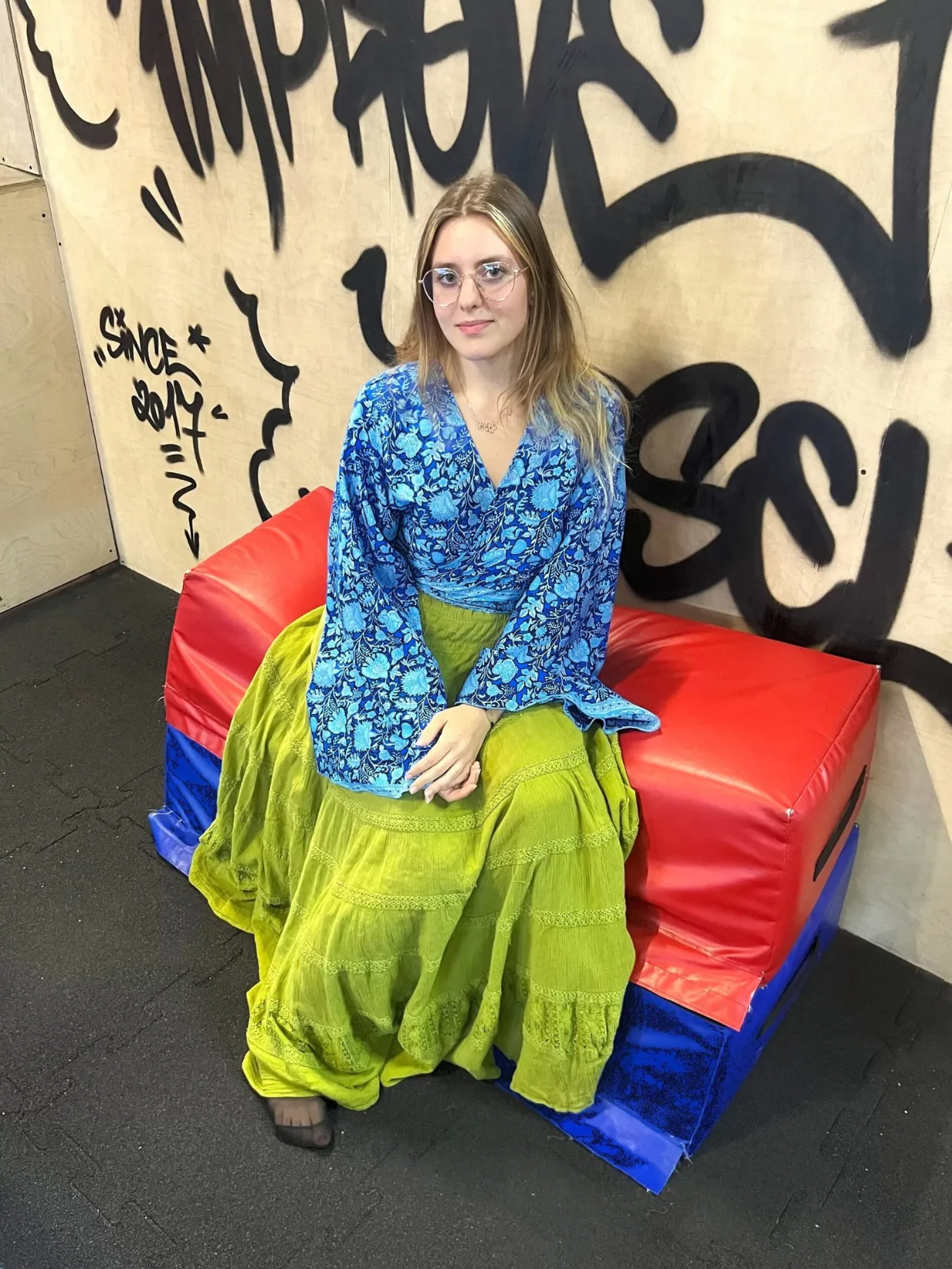 Trenérka Barbora Modrovičová v modrém topu sedí na žíněnce před graffiti stěnou Improve Yourself