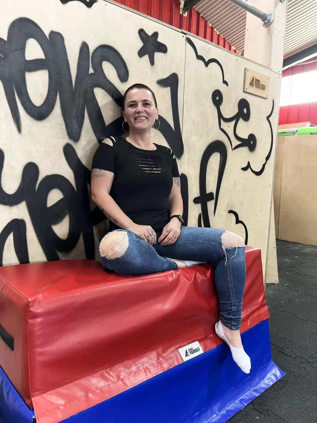 Usměvavá trenérka Ema Onderková sedí na červenomodré žíněnce před graffiti stěnou v parkourové hale