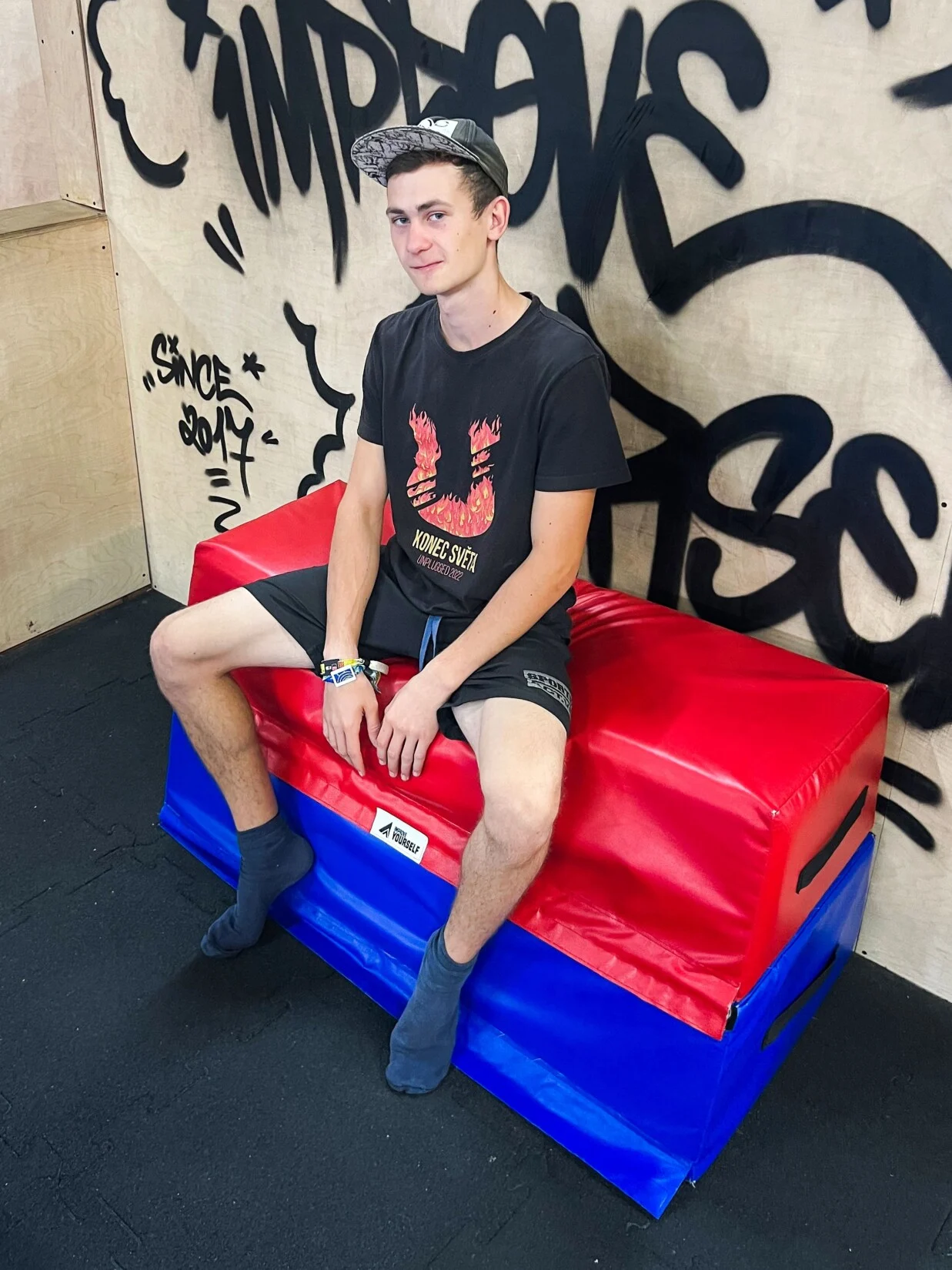 Trenér Jakub Feilhauer s kšiltovkou a černým tričkem sedí na červenomodré žíněnce před graffiti