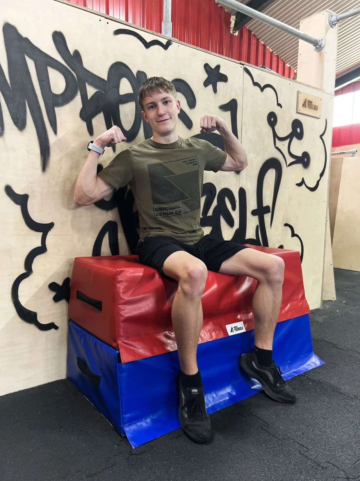 Trenér Jindřich Randa s napjatými bicepsy sedí v khaki tričku na červenomodré žíněnce