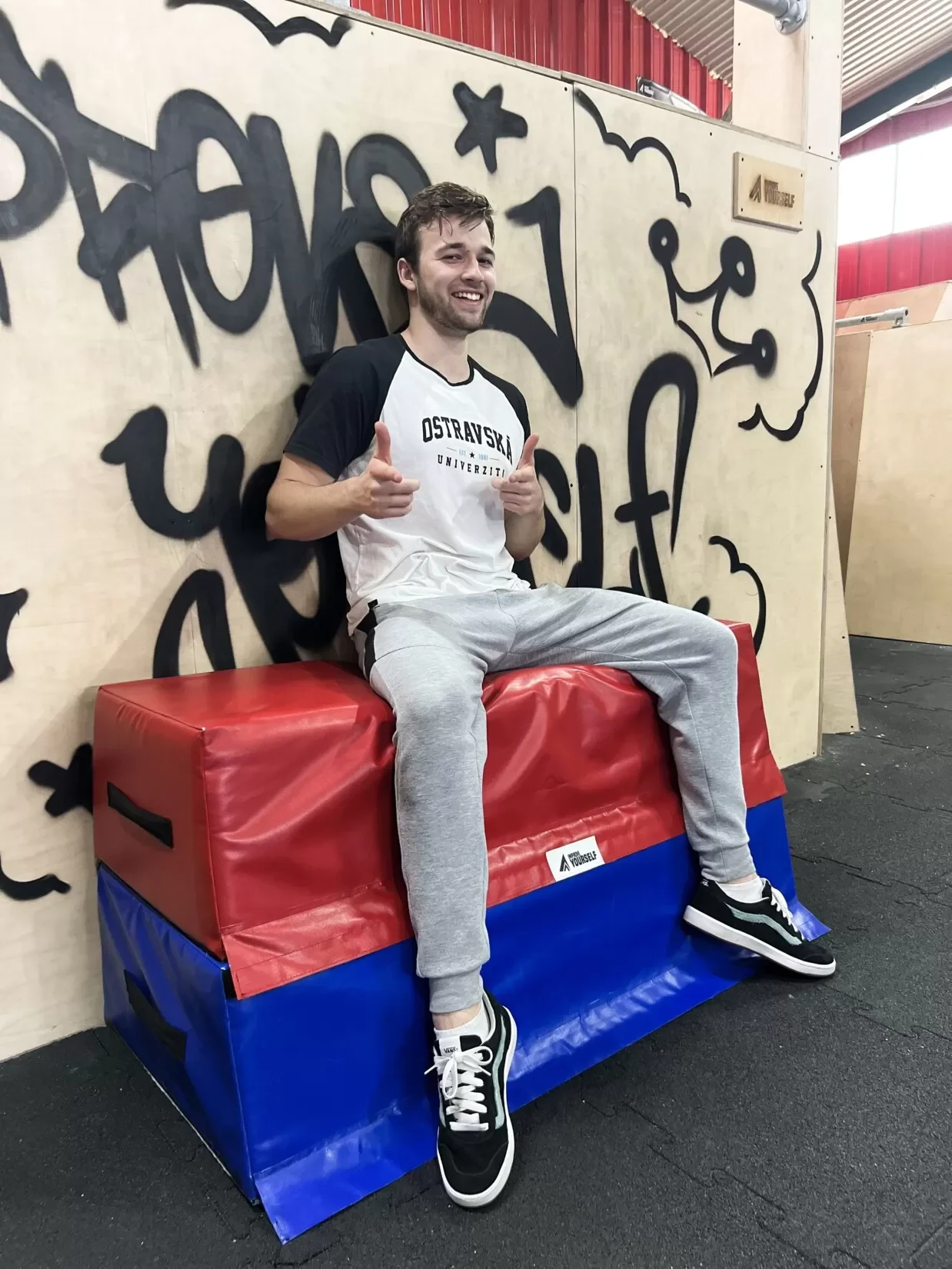 Trenér Jiří Horák s palci nahoru sedí v bílém tričku na červenomodré žíněnce v parkourové hale