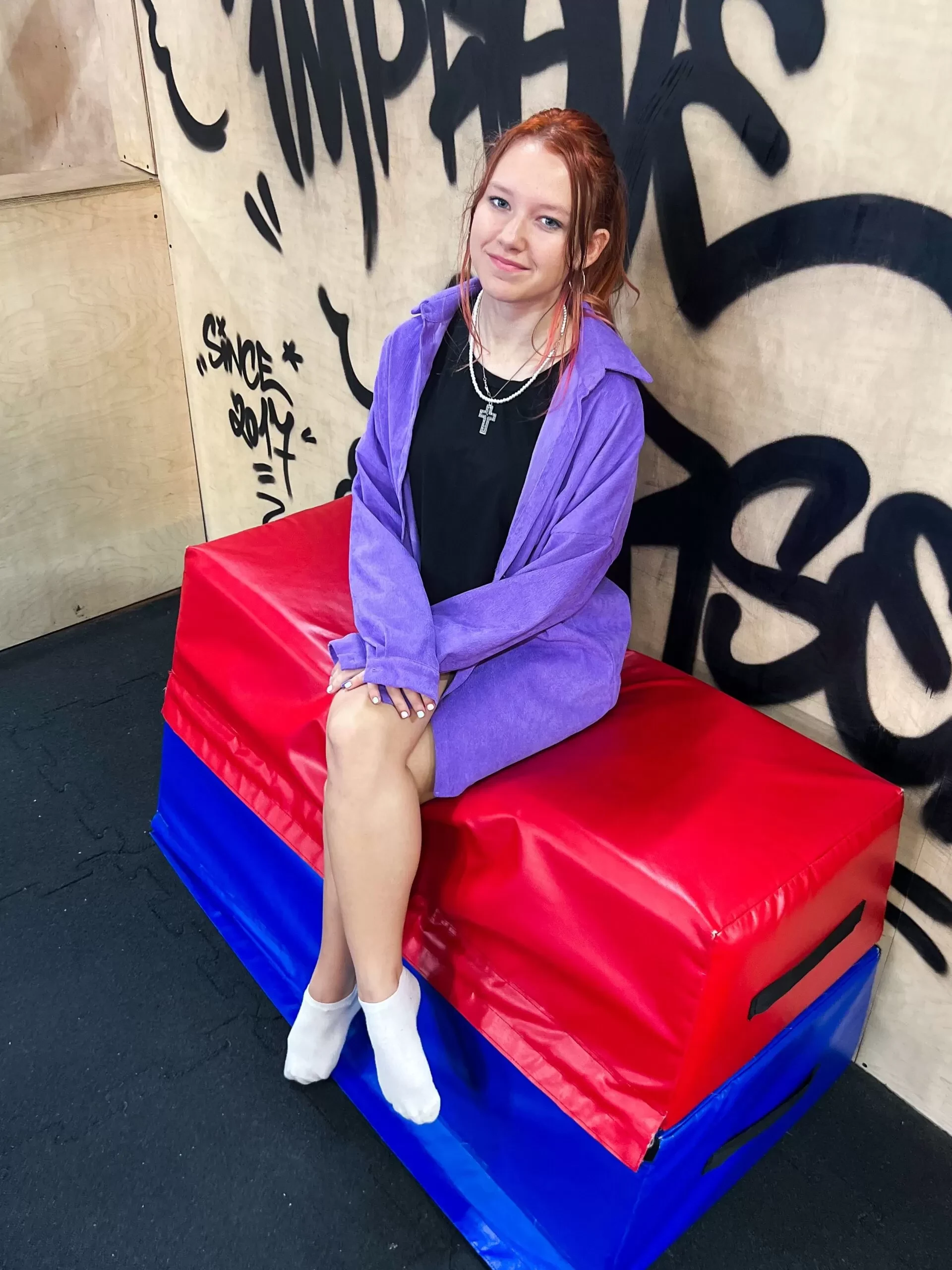 Trenérka Kristýna Ertlová s rusými vlasy a fialovým svetrem sedí na červenomodré žíněnce