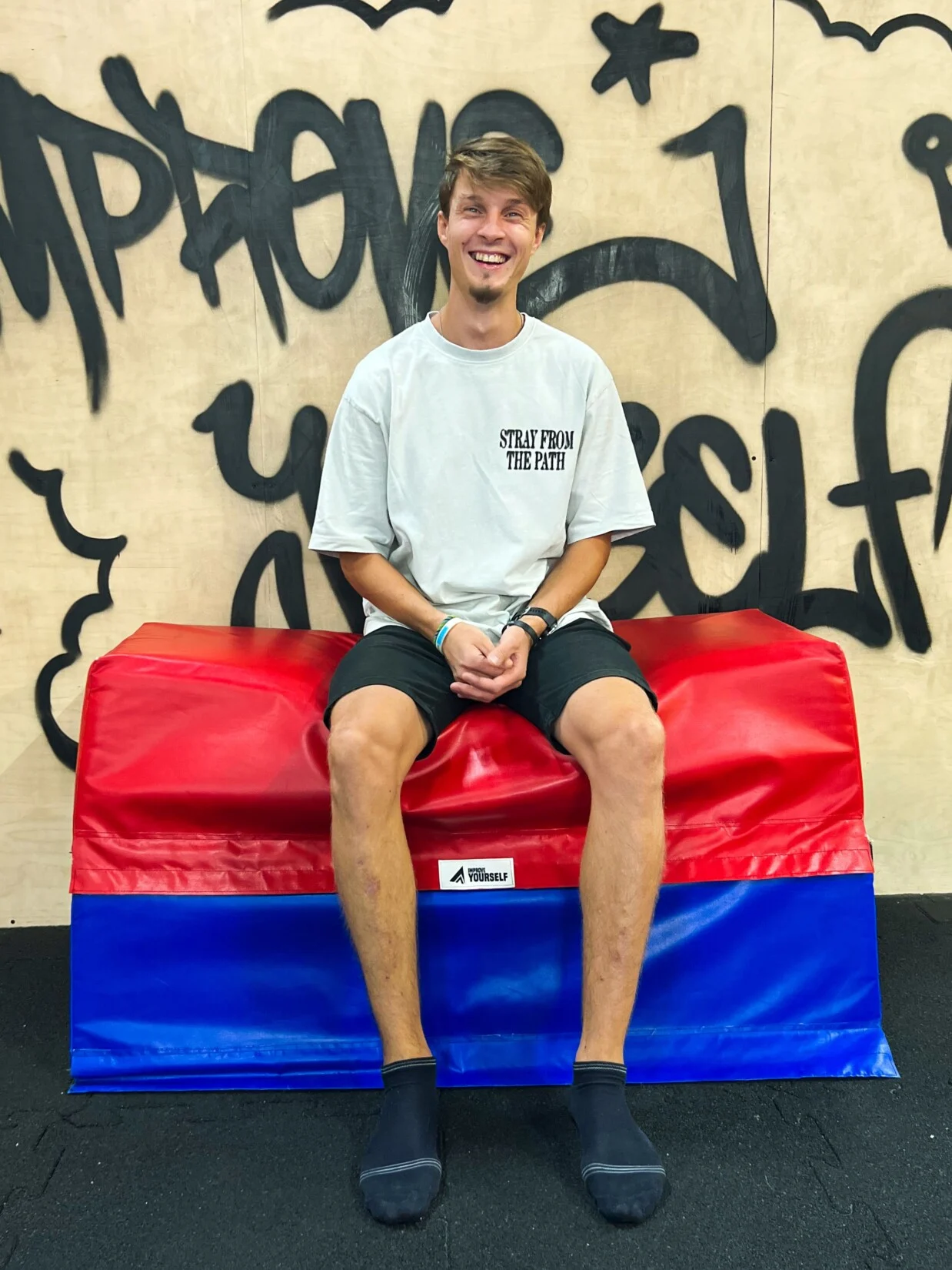 Usměvavý trenér Michal Taláb v bílém tričku sedí na červenomodré žíněnce před graffiti stěnou