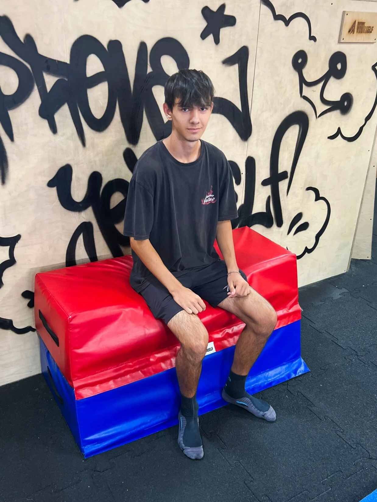 Trenér Oskar Daniheľ v černém tričku sedí na červenomodré žíněnce před graffiti stěnou Improve Yourself