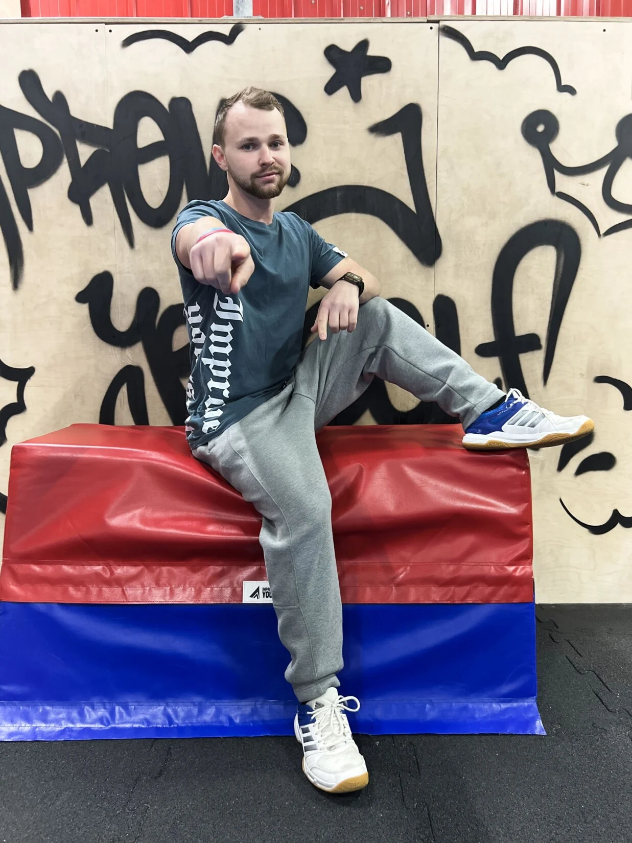 Trenér Petr Bobák v šedém tričku Improve ukazuje prstem do objektivu, sedí na žíněnce