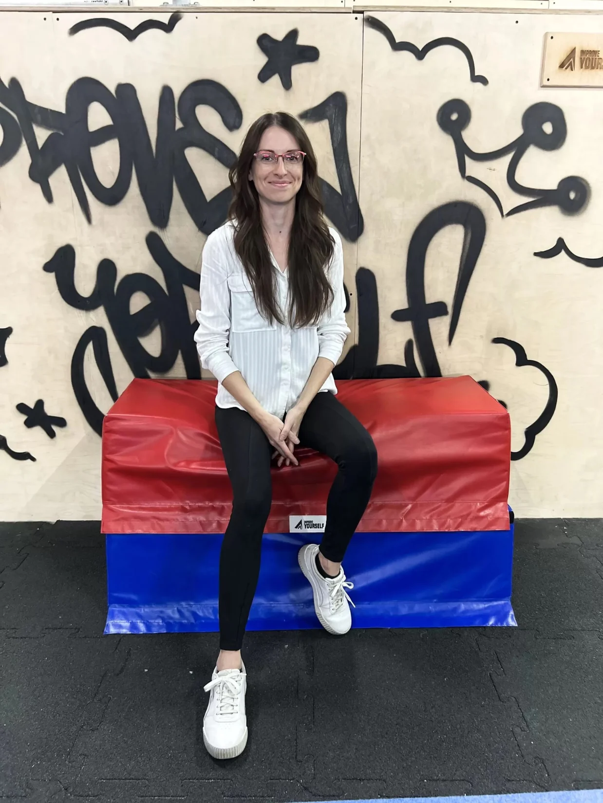 Trenérka Simona Gajdová sedí na červenomodré žíněnce před graffiti stěnou Improve Yourself