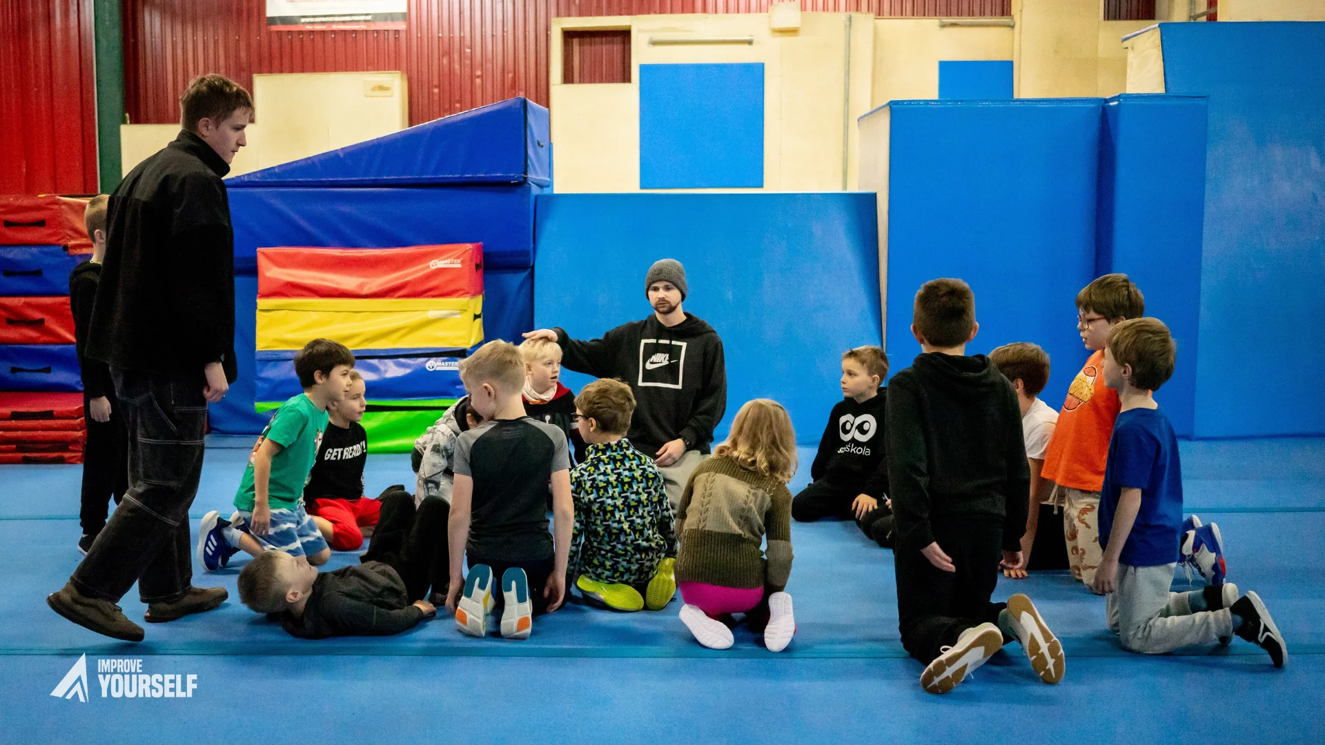 trenér vysvětluje parkour skupině dětí sedících v kruhu na modré žíněnce